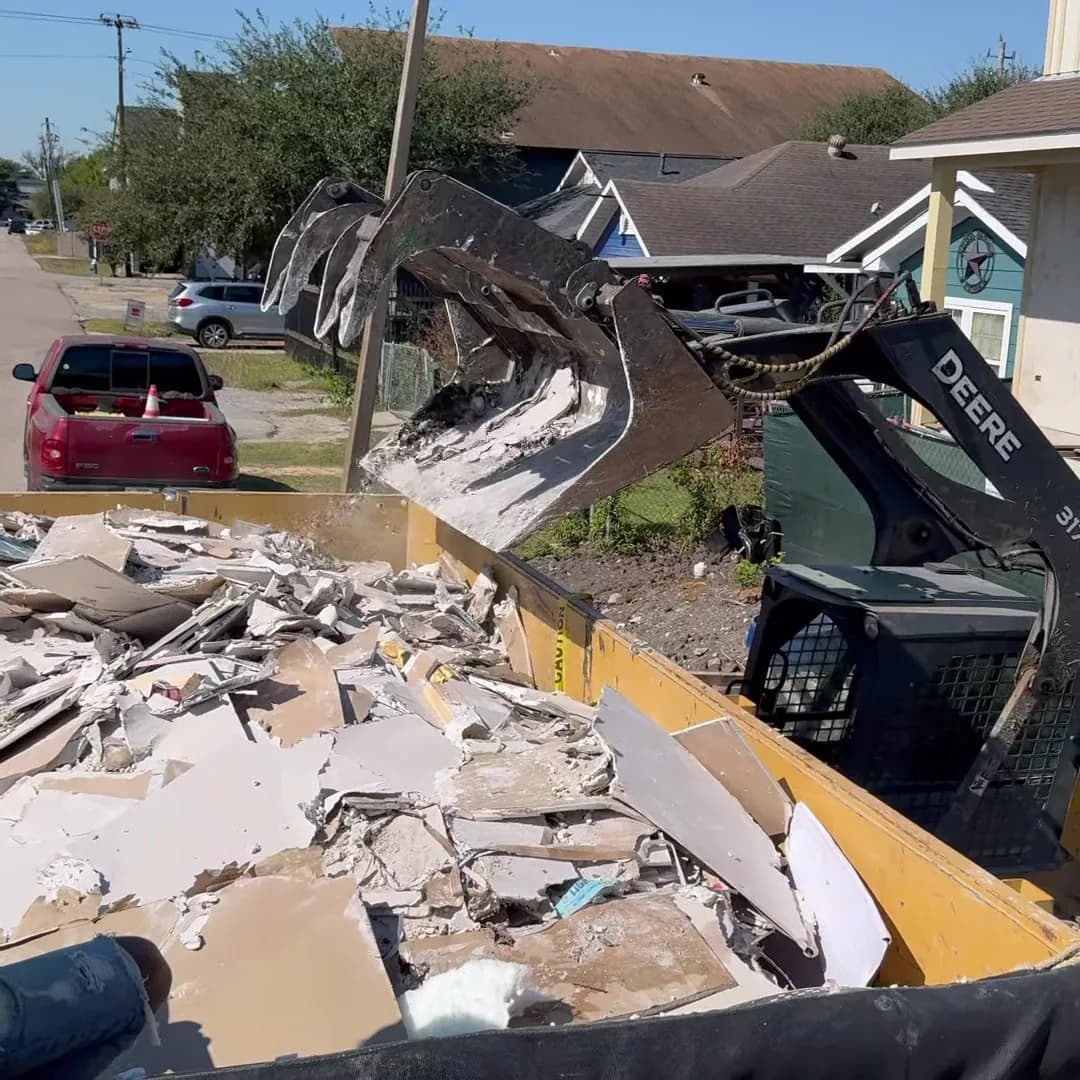 Grapple loading drywall and framing debris into a dump trailer beside a new build